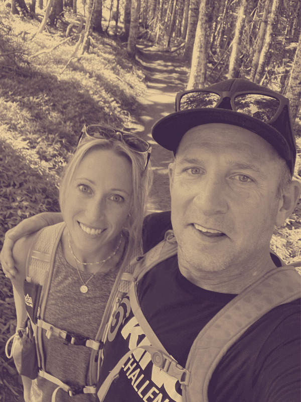 Smiling man and woman in hiking gear take a selfie on a sunny forest trail.