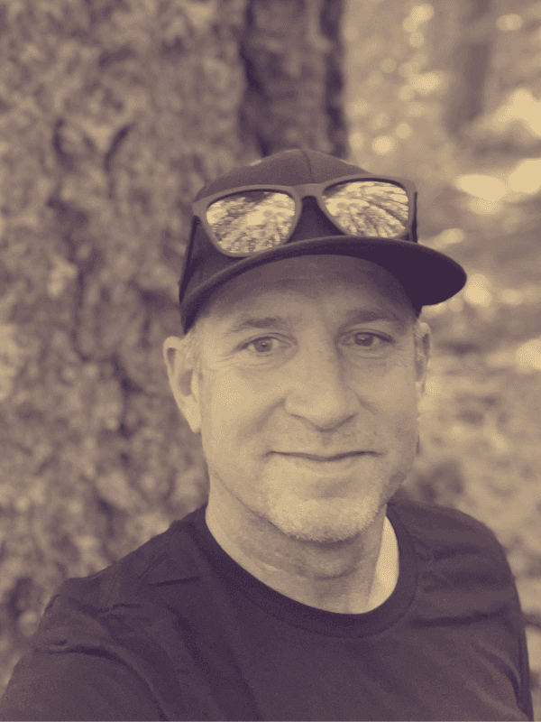 Sepia portrait of a smiling man wearing a black cap and sunglasses against a tree.