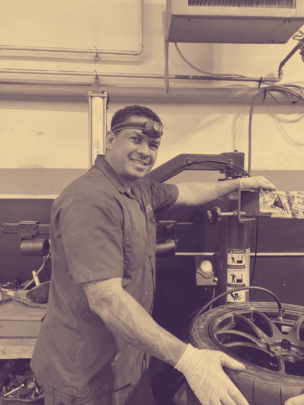 Smiling mechanic wearing a headlamp and gloves operates a tire-changing machine in a garage.